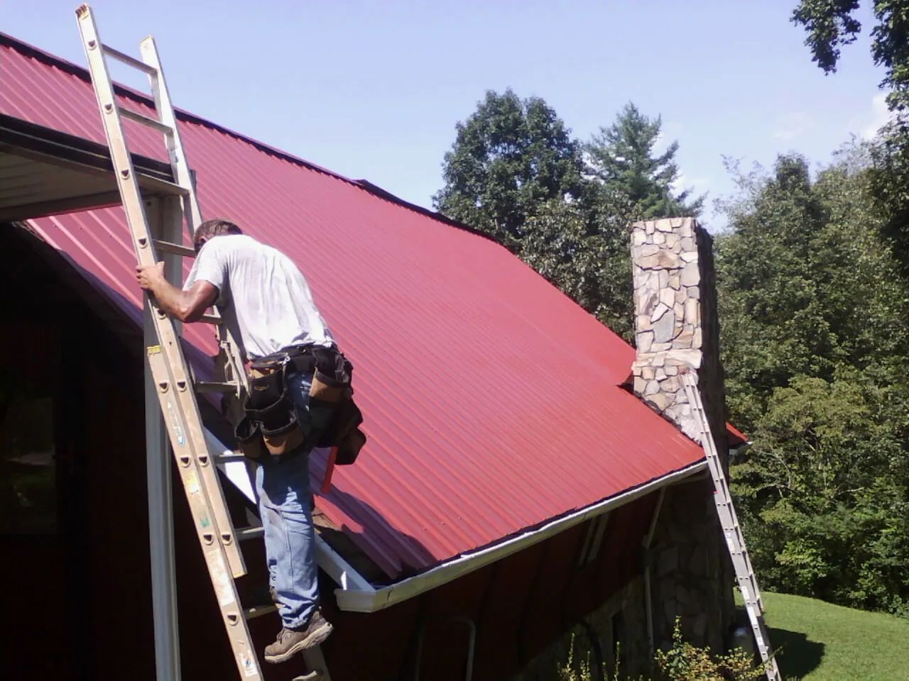 Roofer on a ladder beside a red metal roof and stone chimney — Soffit & Fascia Repair by Roofing Brothers in Sublimity
